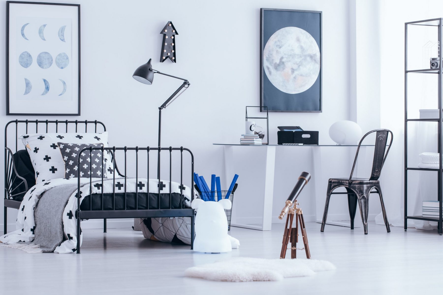 Telescope and black chair at desk in teenager's bedroom interior with metal bed and moon poster on white wall